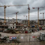 Chantier de construction en activité avec plusieurs grues, briques et matériaux sur le sol; panneaux de sécurité et clôture, paysage urbain en arrière-plan. Problèmes du secteur de la construction évoqué par le contexte.